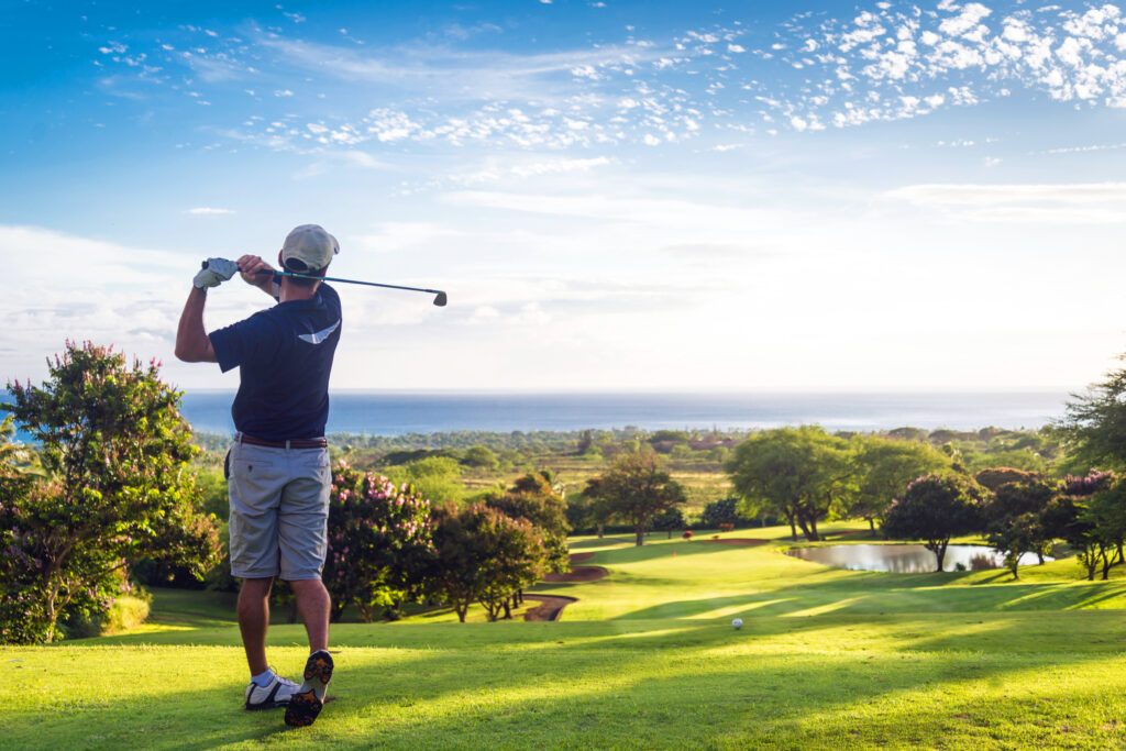 A man in a cap swings a golf club on a sunny fairway overlooking the ocean